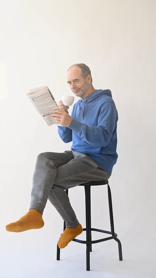 Elderly Man In Blue Hoodie Drinking Coffee And Reading Newspaper 