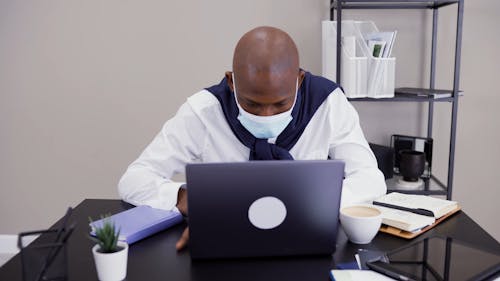 A Man Wearing a Face Mask is Typing on His Laptop While at the Office