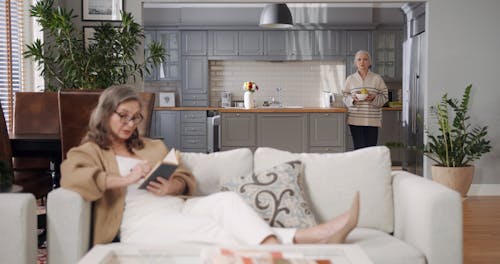 Elderly Women Sitting on Couch while Eating and Reading a Book