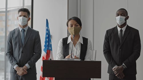 A Woman Speaking on a Podium with Men by Her Side while Wearing Face Masks