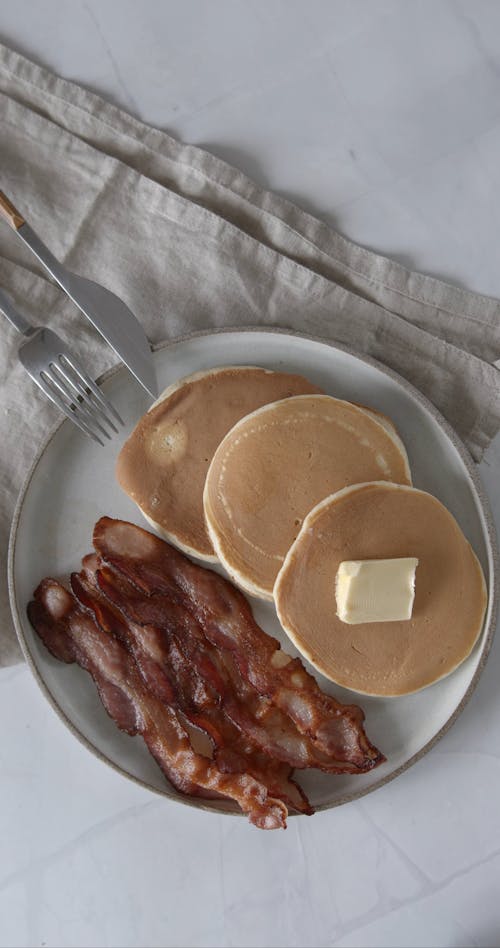 A Person Putting an Egg on a Plate of Bacon and Pancakes