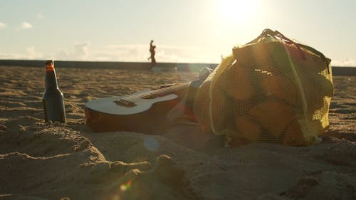 Beer Bottle, Guitar and Bag of Firewood on Beach Sand