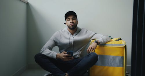 A Delivery Man Sitting Beside a Thermal Bag while Holding a Cellphone