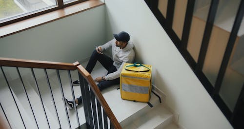 A Delivery Man Sitting Beside a Thermal Bag while Holding a Cellphone