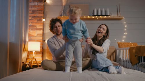 A Boy is Jumping on the Bed While His Parents are Holding Him
