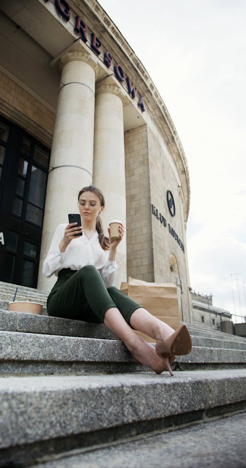 A Woman Using her Cellphone and Drinking Coffee Outside a Building