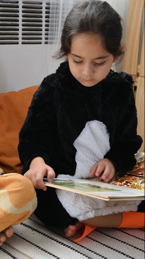 A Little Girl Reading a Book While Sitting on the Floor