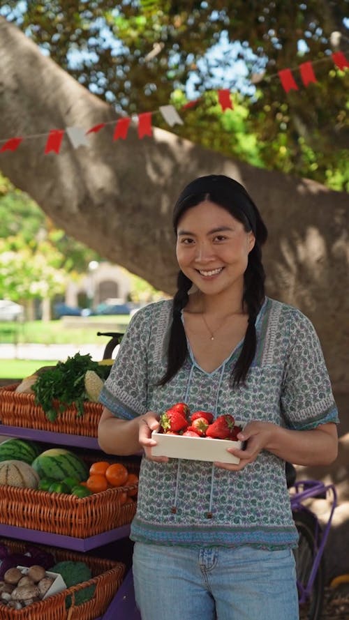 A Woman Smiling while Holding Strawberries beside a Stall