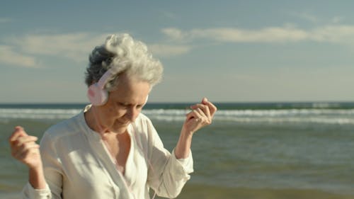 An Elderly Woman Dancing while Using Headphones at the Beach