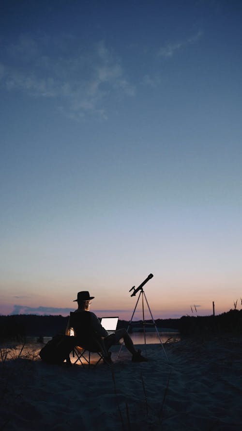 A Man Sitting Outdoors with his Laptop and a Telescope 