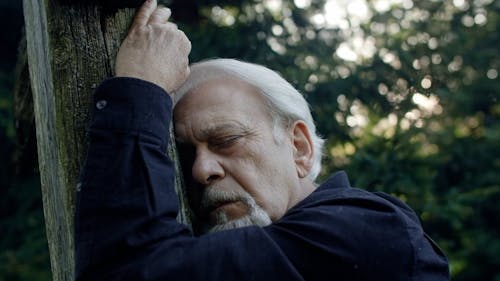 A Sad Elderly Man Leaning on a Wooden Cross